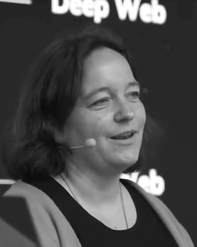 Catrin Schröder-Jaross, Schwarzweissaufnahme auf einer Bühne, Headset-Mikrofon, dunkles Oberteil mit hellem Cardigan, blickt lächelnd zur Seite ins Publikum.